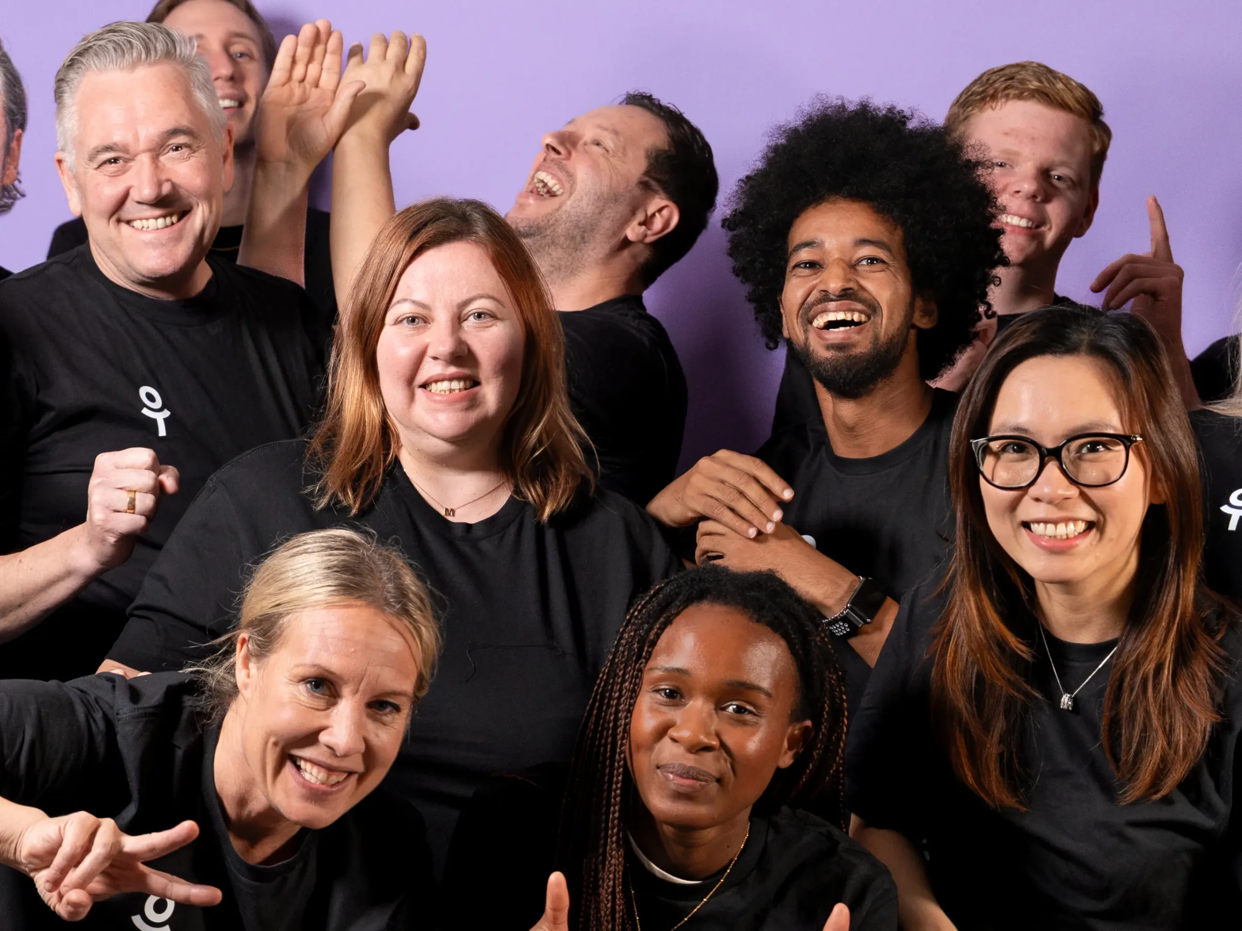 En glad grupp med cirka tio personer i svarta On Track-tröjor skrattar och firar tillsammans framför en lila bakgrund.